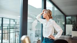 Eine Frau in weißer Bluse und Jeans lehnt an einer Glasscheibe im Office und schaut besorgt.