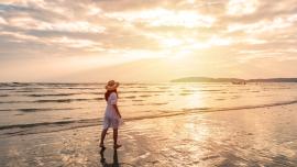 Eine Frau läuft im weißen Kleid bei Sonnenuntergang am Meeresrand durch den nassen Sand.