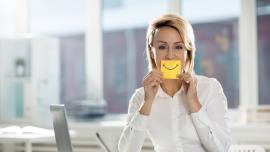 Eine Frau sitzt am Laptop im Büro und hält sich einen gelben Zettel mit aufgemaltem Lächeln vor den Mund.