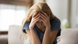 Eine blonde Frau sitzt auf einem Sofa und stützt erschöpft den Kopf in die Hände.