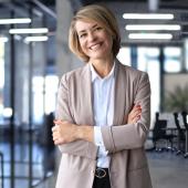 Eine Business-Frau steht mit selbstbewusster Haltung in einem großflächigen Büro und lächelt in die Kamera.