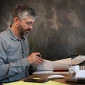 Ein Mann im grauen Businesshemd sitzt konzentriert am Schreibtisch und arbeitet sich durch Dokumente in Papierform.
