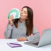 Eine Frau sitzt am Tisch, vor sich Laptop und Notizbuch, in der Hand hält sie aufgefächerte 100-Euro-Scheine, an denen sie mit geschlossenen Augen schnuppert.