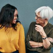 Eine junge schwarze Frau und eine ältere weiße Frau stehen im lockeren Business-Outfit nebeneinander und lachen sich an.