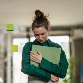 Eine junge Frau in grüner Bluse drückt eine Mappe an sich, sie steht im Büro und schaut unglücklich nach unten