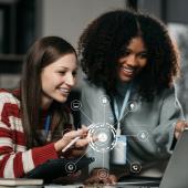 Zwei junge Frauen schauen auf einen Laptop. Symbole für KI, digitale Technik.