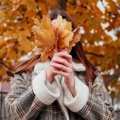 Eine junge Frau im Mantel hält sich Herbstblätter vor das Gesicht, im Hintergrund steht ein Baum mit Herbstlaub