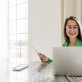 Eine junge Frau sitzt an einem Tisch vor ihrem Laptop und hält ein Tablet in der Hand. Auf dem Tisch liegt ein Smartphone.