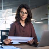 Eine Frau mit dunklen Haaren sitzt im Büro an einem Laptop und schaut auf ein Blatt Papier.