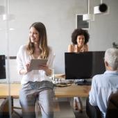 3 Mitarbeitende stehen oder sitzen in einem Büro mit Holztischen und Computerbildschirmen.
