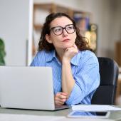 Eine Frau sitzt nachdenklich an einem Laptop und schaut in die Ferne.