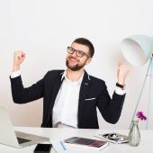 Ein junger Mann im Anzug sitzt an einem Büroschreibtisch und ballt triumphierend die Hände.