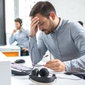 Ein Mann sitzt mit einem anderen in einem Büro am Tisch und stützt müde den Kopf in die Hand.