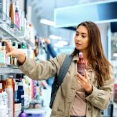 Eine junge Frau in einer Drogerie, sie nimmt eine Tube aus dem Regal und hält eine andere in der Hand.