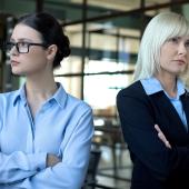 Zwei Frauen in Businesskleidung stehen mit ernsten Gesichtern und verschränkten Armen einander abgewandt.