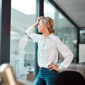 Eine Frau in weißer Bluse und Jeans lehnt an einer Glasscheibe im Office und schaut besorgt.