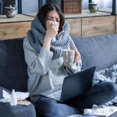 Eine schwarzhaarige Frau sitzt vor einem Laptop auf dem Sofa und putzt sich die Nase, in der Hand hat sie eine Teetasse.