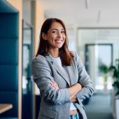 Foto: Drazen - Adobe Stock Eine junge Frau in Blazer steht in einem Büro und lächelt selbstbewusst in die Kamera.