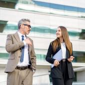 Ein mittelalter Mann und eine junge Frau in Businesskleidung laufen nebeneinander, im Hintergrund sieht man ein Bürogebäude.