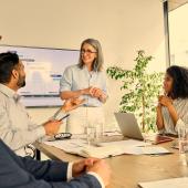 Vier Mitarbeitende sitzen an einem Tisch und schauen zu der mittig stehenden Vorgesetzten, die vor eine Bildschirm steht und einem Mitarbeiter zuhört.
