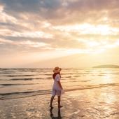 Eine Frau läuft im weißen Kleid bei Sonnenuntergang am Meeresrand durch den nassen Sand.