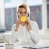 Eine Frau sitzt am Laptop im Büro und hält sich einen gelben Zettel mit aufgemaltem Lächeln vor den Mund.
