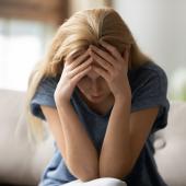 Eine blonde Frau sitzt auf einem Sofa und stützt erschöpft den Kopf in die Hände.