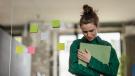 Eine junge Frau in grüner Bluse drückt eine Mappe an sich, sie steht im Büro und schaut unglücklich nach unten
