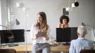 3 Mitarbeitende stehen oder sitzen in einem Büro mit Holztischen und Computerbildschirmen.