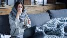 Eine schwarzhaarige Frau sitzt vor einem Laptop auf dem Sofa und putzt sich die Nase, in der Hand hat sie eine Teetasse.