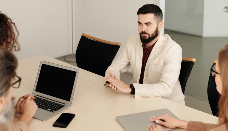 Vier Mitarbeitende sitzen am Tisch, drei davon haben bereits Arbeitsmaterialien, der Neue noch nicht.