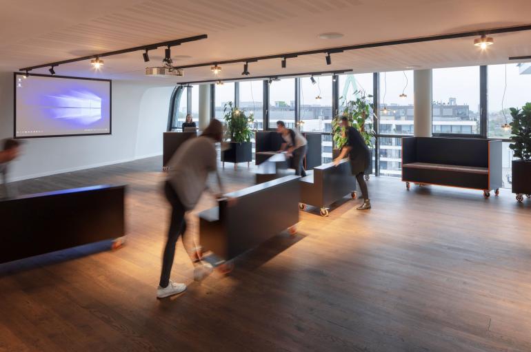 Kommunikationslab bei Microsoft. Großer Raum, Mitarbeitende räumen Sofas um.