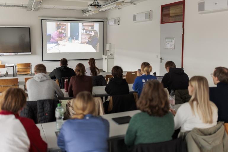 Studierende blicken auf eine Leinwand, auf der die Gesprächssimulation zu sehen ist.