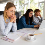 Eine Frau sitzt still und nachdenklich vor ihren Unterlagen. Neben ihr am Tisch sitzen zwei Kolleg*innen.