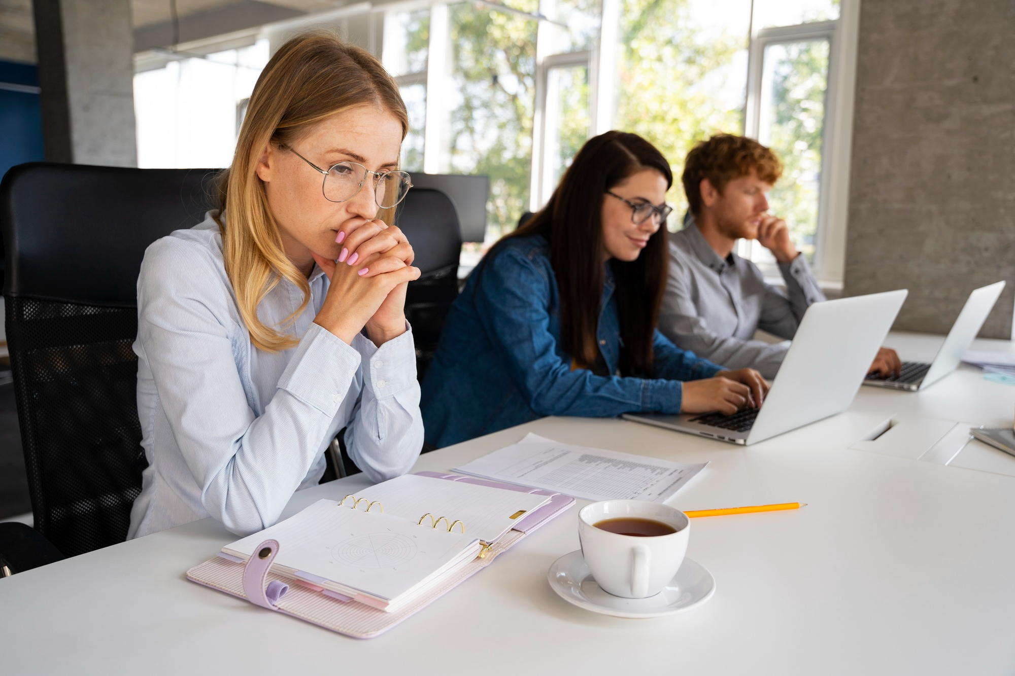 Eine Frau sitzt still und nachdenklich vor ihren Unterlagen. Neben ihr am Tisch sitzen zwei Kolleg*innen.