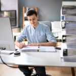 Ein Mann sitzt neben einem Stapel Aktenordner am Bürotisch