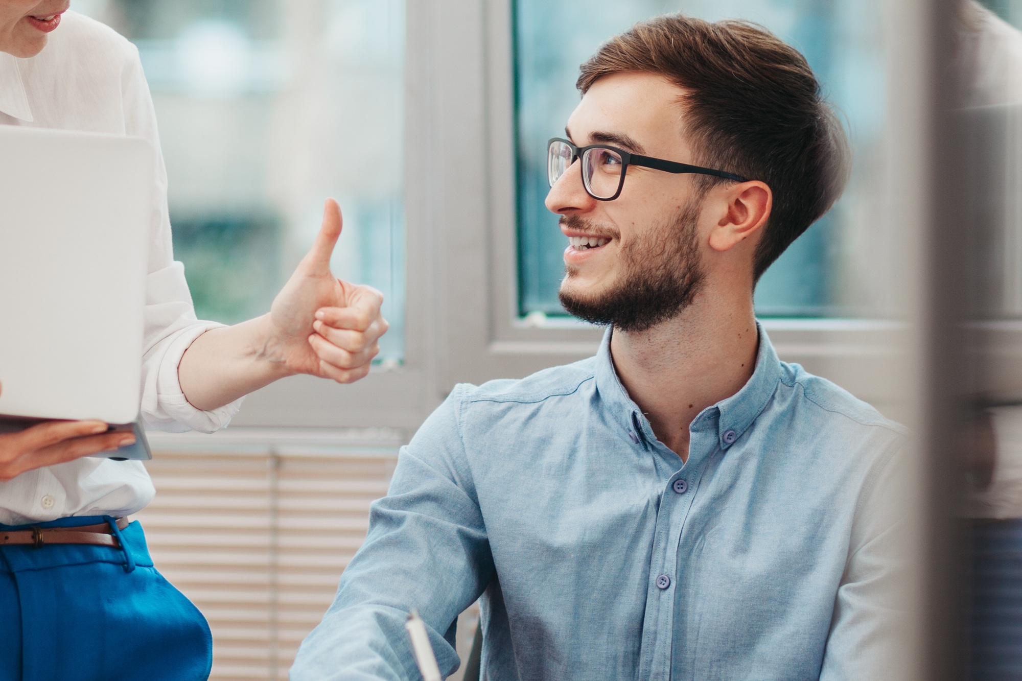 Ein junger Mann sitzt am Tisch, neben ihm steht seine Vorgesetzte und zeigt ihm einen Daumen hoch.