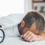 Ein Mann stützt das Gesicht in die Hand und schaltet den Wecker aus. Er liegt noch im Bett.
