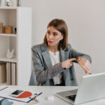 Eine Frau sitzt im Büro und tippt vorwurfsvoll auf ihre Uhr.
